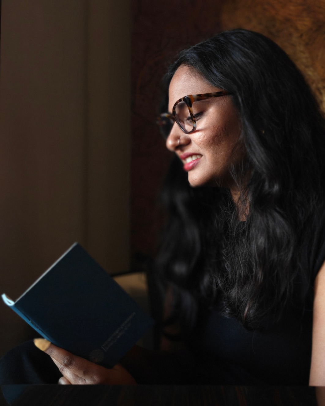 A woman with long dark hair and tortoiseshell glasses is shown in profile, smiling while holding a dark blue book in a warmly lit indoor setting.